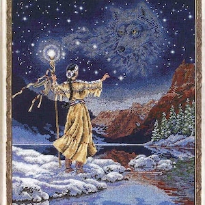 May include: A framed cross-stitch embroidery depicting a figure in tan fringed clothing holding a glowing staff, standing on a snowy bank by a lake. A large spectral wolf head appears in the starry night sky above mountains and evergreen trees.