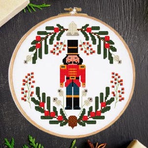 May include: A cross-stitch pattern featuring a traditional Christmas nutcracker standing in the center of a wreath of holly and berries.