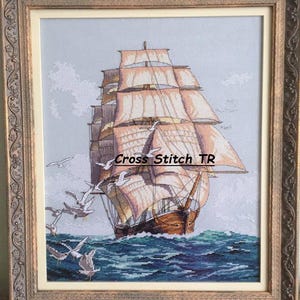 Könnte beinhalten: Gerahmtes Kreuzstich-Kunstwerk, das ein Segelschiff mit beigen Segeln und einem braunen Rumpf auf blauem Meer zeigt. Weiße Möwen fliegen im Vordergrund. Der Rahmen ist goldfarben und verziert. Der Text "Cross Stitch TR" ist im Bild zu sehen.