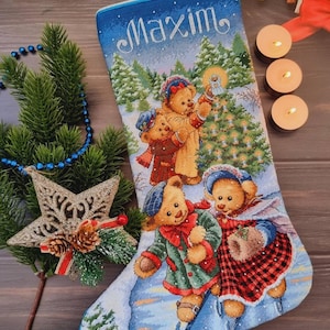 May include: Embroidered Christmas stocking with a winter scene featuring teddy bears ice skating and carrying a lantern. The name "Maxim" is embroidered in cursive at the top. The stocking is blue and white with a snowy landscape.