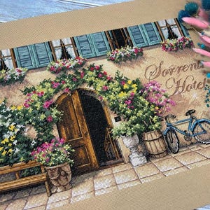 May include: Cross-stitch of the Sorrento Hotel facade, featuring a wooden door framed by flowers. A blue bicycle rests nearby. The building has blue shutters and window boxes filled with flowers. The text "Sorrento Hotel" is visible.