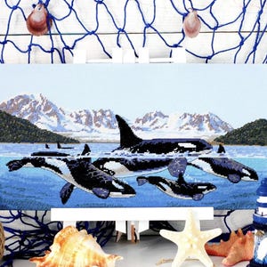 May include: A cross-stitch artwork depicting a pod of orcas swimming in the ocean. The black and white whales are set against a backdrop of snow-capped mountains and a blue sky. The artwork is displayed on a white easel.