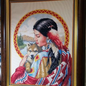 May include: A framed cross-stitch embroidery depicting a person with dark hair and a feather accessory holding a small wolf cub. The scene is set against a circular background featuring a landscape with mountains and water, all enclosed in a dark wood frame.