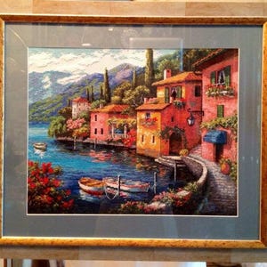 May include: Framed cross-stitch artwork showing a waterfront village scene. The artwork features colourful buildings, boats, and lush greenery along a blue lake. The frame is a light brown colour.