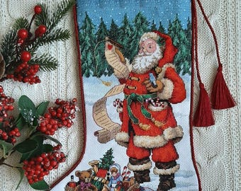 May include: Embroidered Christmas stocking depicting Santa Claus in a red suit, holding a list and gifts. The stocking has a blue background with a snowy scene and a bag of toys. Red tassels and a hanging loop complete the design.
