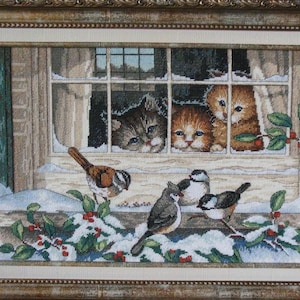 Puede incluir: Arte de bordado enmarcado que representa una escena invernal. Tres gatitos miran por una ventana a pájaros posados en ramas cubiertas de nieve con bayas rojas. La obra de arte está en un marco dorado.