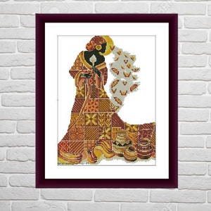 May include: Cross-stitch art of a figure in a patterned dress, holding a candle, with butterflies and pottery. The artwork is framed in a dark burgundy frame against a white brick wall. The color palette includes shades of brown, gold, and red.