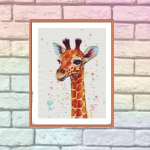 Op de afbeelding: Een ingelijst kruissteekpatroon van een giraffe met een witte achtergrond en kleurrijke verfspatten. De giraffe is bruin en oranje met een lange nek en grote oren.