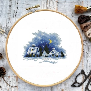 Könnte beinhalten: Ein Kreuzstichmuster, das eine verschneite Winterlandschaft mit drei Häusern, Bäumen und einem Halbmond am Himmel zeigt. Die Häuser haben gelbe Fenster und sind mit Schnee bedeckt. Die Bäume sind grün und der Himmel ist blau mit weißen Sternen.
