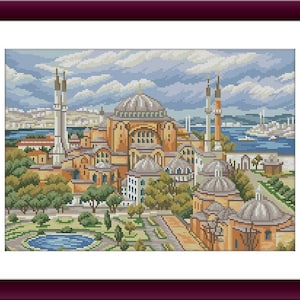 May include: A framed cross-stitch depicting a detailed cityscape with a large domed building, minarets, and other structures. The artwork features a color palette of beige, orange, green, and blue, with a body of water and a cloudy sky in the background.