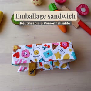 Può includere: Un involucro per sandwich riutilizzabile con sfondo bianco e motivi colorati di cibo e oggetti. L'involucro ha un'etichetta gialla e il testo "Emballage sandwich Réutilisable & Personnalisable".