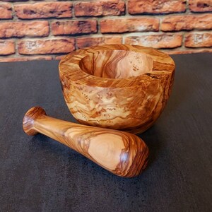May include: A wooden mortar and pestle set made from olive wood. The mortar is a deep bowl with a natural wood grain pattern. The pestle is a smooth, rounded wooden stick.