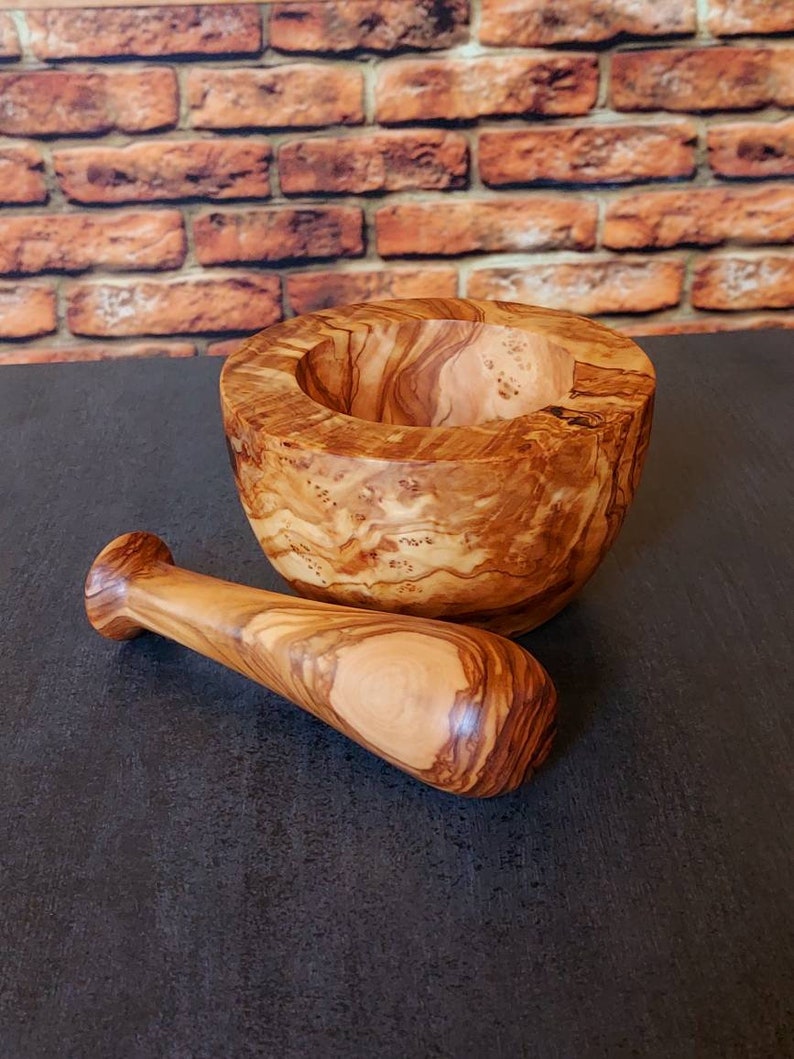 May include: A wooden mortar and pestle set made from olive wood. The mortar is a deep bowl with a natural wood grain pattern. The pestle is a smooth, rounded wooden stick.
