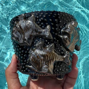 Peut inclure: Un pot de fleurs cylindrique en céramique avec une surface sombre, texturée et bosselée, orné de trois sculptures de poissons en relief aux tons métalliques. Le récipient repose sur de petits pieds intégrés à la base, avec une finition brillante.
