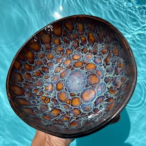 May include: A round ceramic bowl featuring a complex, multi-colored glaze with a honeycomb-like pattern. The interior surface displays a vibrant mix of deep amber, metallic blue, and iridescent purple tones, creating a textured, crystalline appearance.