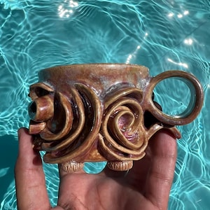 May include: A ceramic mug featuring a glossy, multi-toned brown and reddish glaze with prominent, raised spiral relief patterns on the exterior. The mug includes a circular handle and rests on two small, textured feet at the base of the vessel.
