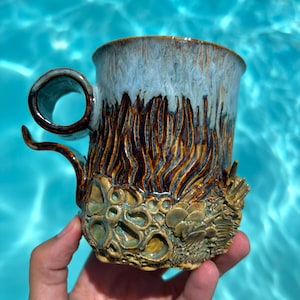 May include: A ceramic mug featuring a light blue glazed rim that transitions into a dark brown, textured body. The lower section displays intricate, raised organic shapes in shades of green and tan, resembling coral or fungal growth.