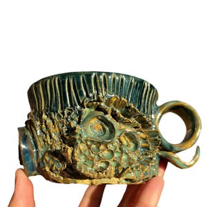 Pode incluir: Uma caneca de cerâmica artesanal com um design texturizado único. A caneca apresenta um esquema de cores verde-azulado e dourado com um padrão orgânico esculpido. A alça é um laço curvo e a caneca parece ser pintada à mão.