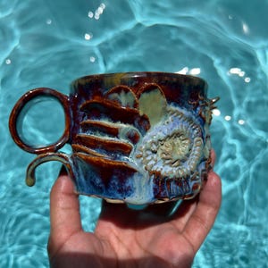 May include: A ceramic mug featuring a glossy, multi-colored glaze in shades of deep blue, brown, and cream. The exterior surface is decorated with raised, textured organic shapes and a circular, fossil-like relief pattern on the side.