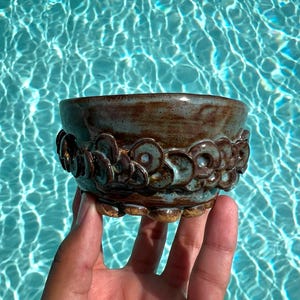 May include: A small, round ceramic bowl featuring a glossy, mottled brown and teal glaze. The exterior is decorated with a textured, repeating pattern of overlapping circular clay discs that wrap around the middle and base of the vessel.