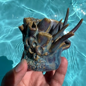 May include: A ceramic mug featuring a textured, organic surface with protruding, horn-like structures and deep, cratered openings. The glaze displays a marbled blend of deep blue, purple, and earthy brown tones across the entire exterior surface.
