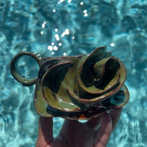 May include: A handmade ceramic mug with a unique, sculpted design. The mug features a dark green glaze with brown accents, and a distinctive, swirling shape. The handle is a simple loop. The mug is held against a background of blue water.