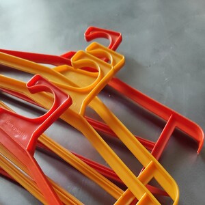 May include: A stack of orange and red plastic hangers. The hangers are made of a thin, flexible plastic and have a simple design. The text "dem plastik" is printed on one of the hangers.