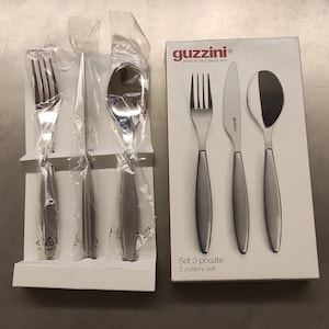 May include: A set of stainless steel cutlery, including a fork, knife, and spoon, packaged in clear plastic. A boxed set of three pieces of cutlery is also visible, with the brand name "guzzini" and the text "Set 3 posate 3 cutlery set".