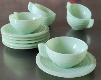 May include: A collection of vintage jadeite green teacups and saucers. The cups have handles and sit on matching saucers. The set includes stacked saucers and several cups, all with a ribbed design. The tableware is arranged on a reflective surface.