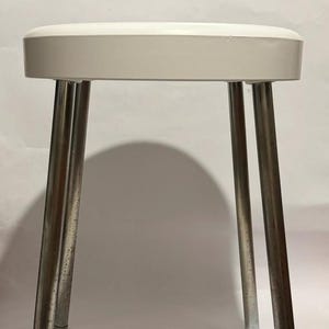 Peut inclure: Un tabouret en plastique blanc avec un siège rond et trois pieds chromés.