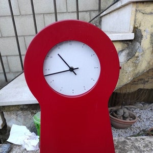 May include: A red clock with a white face and black hands. The clock is shaped like a keyhole and is sitting on a stone surface.