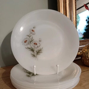 Puede incluir: Una pila de cinco platos de cena de cerámica blanca con un diseño floral con margaritas blancas y hojas verdes. Los platos están apilados sobre una superficie de madera.