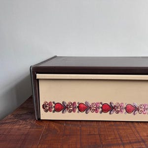 Puede incluir: Una caja de pan de metal marrón y beige con un diseño floral con peras rojas. La caja de pan tiene un cajón extraíble y el texto "Brabantia" está impreso en la parte delantera.