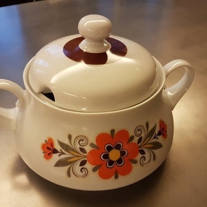 Op de afbeelding: Witte keramische serveerschaal met deksel. De schaal heeft een bloemenmotief met oranje, rode en bruine bloemen. Het deksel heeft een bruine cirkel in het midden.