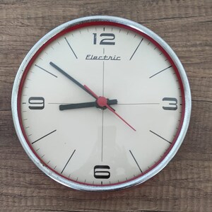 Op de afbeelding: Een witte analoge klok met zwarte wijzers en rode accenten. De wijzerplaat van de klok heeft zwarte cijfers en de woorden "Electric" in rood.