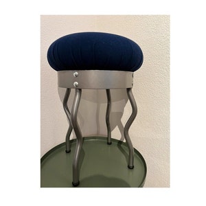 May include: A blue upholstered stool with a metal base and four wavy legs. The stool is sitting on a green surface.