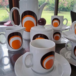 Puede incluir: Un juego de café de cerámica blanca con un diseño retro. El juego incluye una cafetera, una lechera, un azucarero y varias tazas y platillos. Cada pieza presenta un patrón circular marrón, blanco y naranja. El juego se exhibe sobre una superficie reflectante.