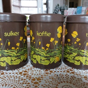 Puede incluir: Tres botes de metal marrón con diseños de flores amarillas. Los botes están etiquetados como "suiker", "koffie" y "thee".