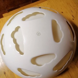 May include: A white, spherical decorative object with cloud-shaped cutouts. The object has a handle and a small, circular indentation on the top. The surface is smooth and the overall design is modern and minimalist.