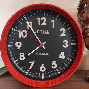 May include: A red and black round clock with white numbers and hands. The clock face says "LOWELL electronic".
