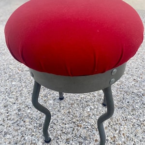 May include: A red upholstered stool with a round seat and three curved metal legs. The stool is made of a sturdy metal frame and has a soft, plush seat.