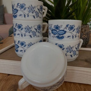 Può includere: Un set di quattro tazze in ceramica bianca con un motivo floreale blu. Le tazze sono impilate l'una sull'altra, con la tazza superiore capovolta.