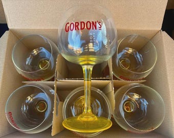 Könnte beinhalten: Ein Set aus sechs transparenten Gin-Gläsern mit gelbem Fuß und Stiel und dem roten "Gordon's"-Logo. Die Gläser sind in einem Karton angeordnet, wobei ein Glas aufrecht und die anderen umgedreht sind.
