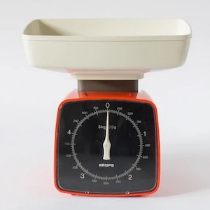 Puede incluir: Una b&aacute;scula de cocina roja y blanca con un dial negro y una bandeja de pesaje blanca. El dial indica "5kg 25g" y tiene marcas para gramos y kilogramos. La b&aacute;scula est&aacute; fabricada por Krups.