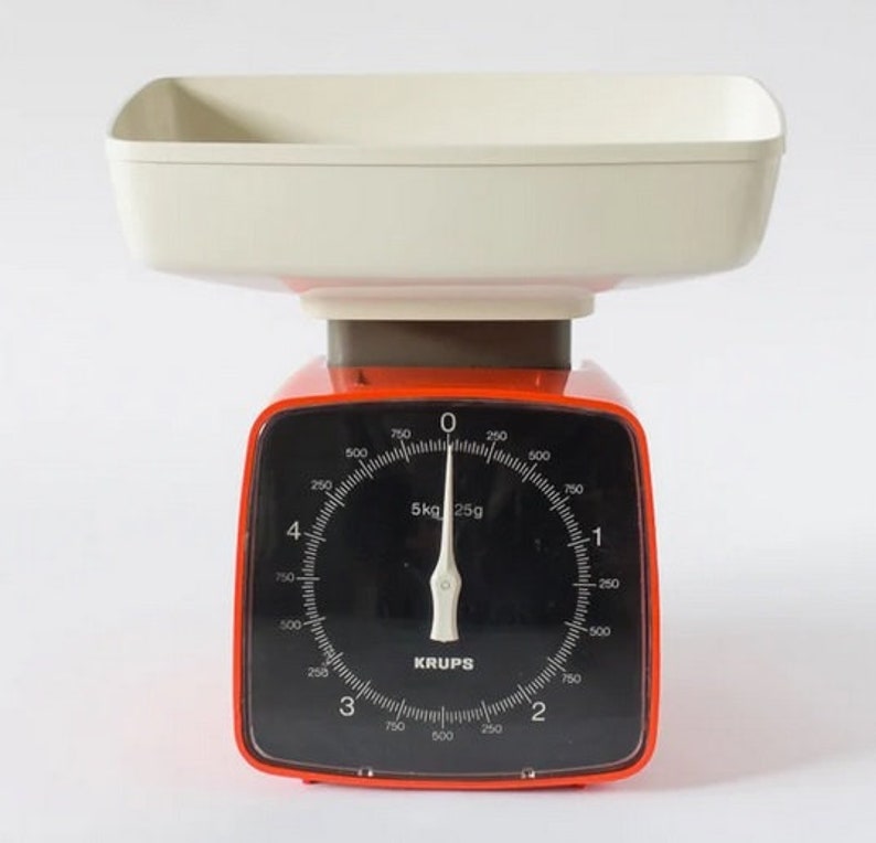 Puede incluir: Una b&aacute;scula de cocina roja y blanca con un dial negro y una bandeja de pesaje blanca. El dial indica "5kg 25g" y tiene marcas para gramos y kilogramos. La b&aacute;scula est&aacute; fabricada por Krups.