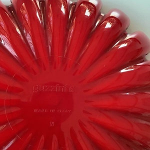 K&ouml;nnte beinhalten: Eine leuchtend rote, bl&uuml;tenf&ouml;rmige Servierschale. Die Schale hat eine gl&auml;nzende Oberfl&auml;che und ein geriffeltes Design, mit dem Text "guzzini" und "MADE IN ITALY" sichtbar. Die Schale ist wahrscheinlich aus Kunststoff oder einem &auml;hnlichen Material.