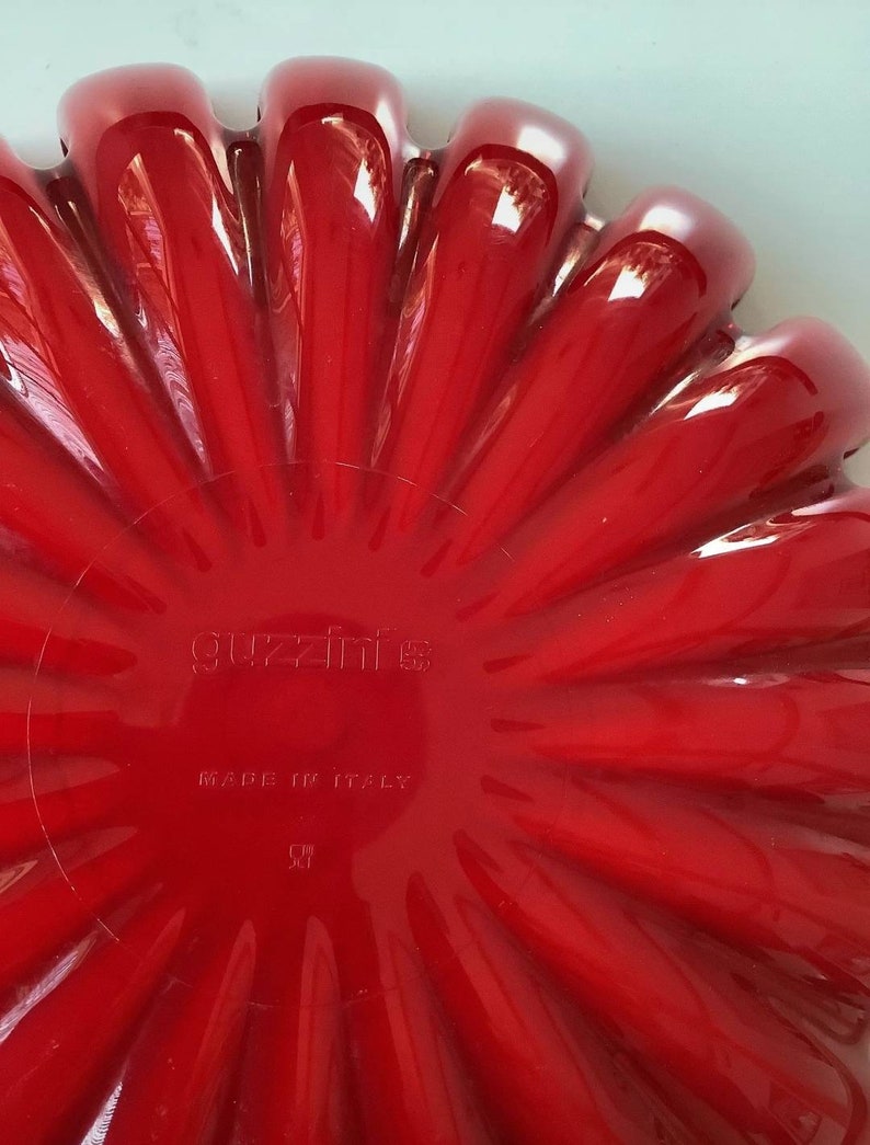 K&ouml;nnte beinhalten: Eine leuchtend rote, bl&uuml;tenf&ouml;rmige Servierschale. Die Schale hat eine gl&auml;nzende Oberfl&auml;che und ein geriffeltes Design, mit dem Text "guzzini" und "MADE IN ITALY" sichtbar. Die Schale ist wahrscheinlich aus Kunststoff oder einem &auml;hnlichen Material.