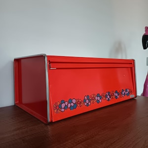 Op de afbeelding: Een rode metalen broodtrommel met een zilveren rand. De broodtrommel heeft een bloemenontwerp in blauw, roze en paars langs de onderrand. De broodtrommel is rechthoekig en staat op een houten oppervlak.