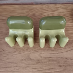 Puede incluir: Dos herramientas de jardín de cerámica con forma de mano. Cada herramienta tiene una base verde claro con cuatro púas en forma de dedo y una parte superior rectangular verde oscuro. Las herramientas están diseñadas para jardinería y están sobre una superficie de madera.