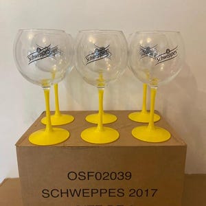 Puede incluir: Seis copas transparentes en forma de globo con tallos y bases amarillas, cada una con el logotipo de Schweppes. Las copas están sobre una caja de cartón con el texto "OSF02039 SCHWEPPES 2017 BOITE DE 6".